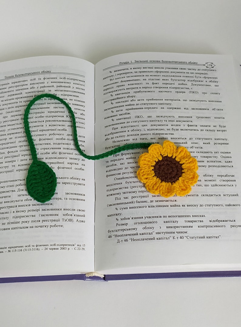 May include: A crocheted sunflower bookmark with a green stem and leaves. The bookmark is laying on top of an open book with text about accounting.