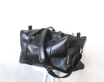 May include: A black leather duffel bag with a top handle and a shoulder strap. The bag is sitting on a white stool with wooden legs.