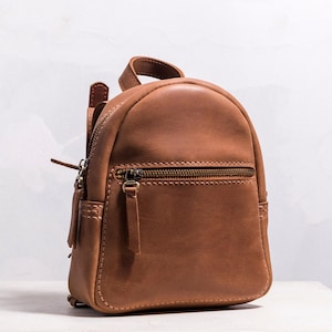 May include: A small, brown leather backpack with a rounded top and a front zippered pocket. The bag has a top handle and adjustable straps. The leather has a natural grain and a matte finish. The hardware is a dark bronze color.
