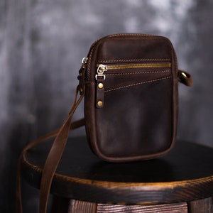 May include: A brown leather crossbody bag with a zipper closure and gold hardware. The bag is sitting on a wooden stool.