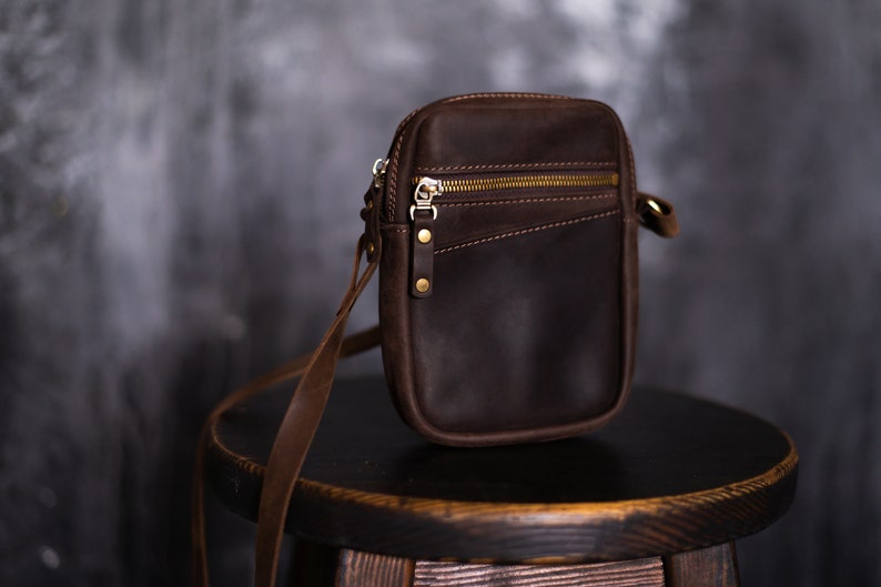 May include: A brown leather crossbody bag with a zipper closure and gold hardware. The bag is sitting on a wooden stool.
