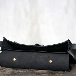 May include: A black leather bag with a curved top and a long strap. The bag has a rectangular body with two brass-coloured rivets. The bag is open, revealing the interior.