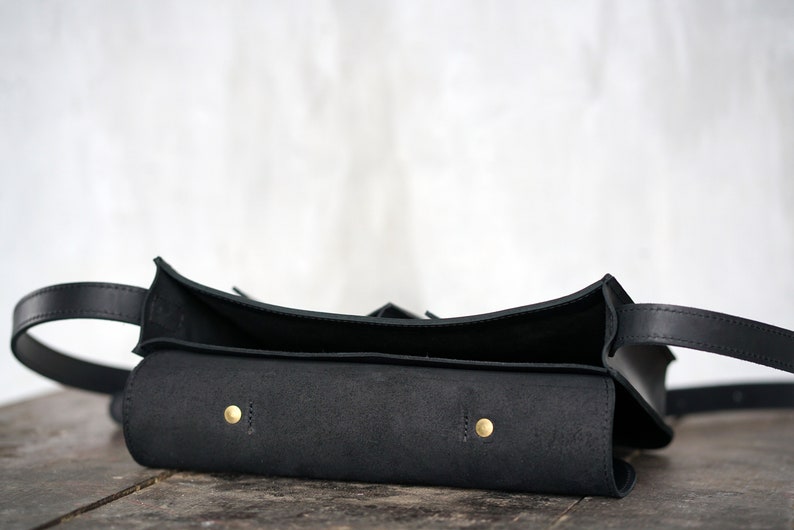 May include: A black leather bag with a curved top and a long strap. The bag has a rectangular body with two brass-coloured rivets. The bag is open, revealing the interior.