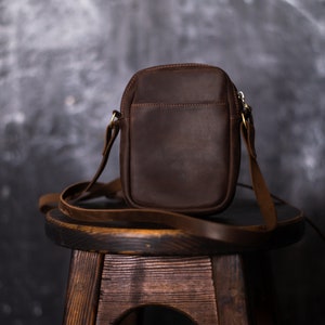 May include: A brown leather crossbody bag with a zipper closure and a long adjustable strap. The bag is sitting on a wooden stool.