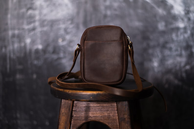 May include: A brown leather crossbody bag with a zipper closure and a long adjustable strap. The bag is sitting on a wooden stool.