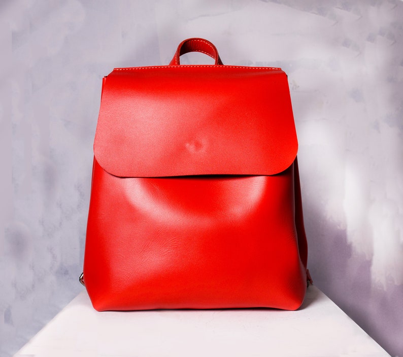 May include: A bright red leather backpack with a flap closure. The backpack has a simple design and is made of high-quality leather.