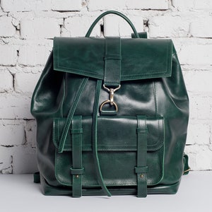 May include: A dark green leather backpack with multiple pockets and straps. The backpack has a flap closure with a gold buckle.