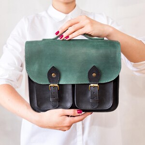 May include: A two-tone leather satchel with a green flap and black body. The bag features two front pockets, gold-coloured buckles, and a top handle. The satchel is held up by a person wearing a white shirt.