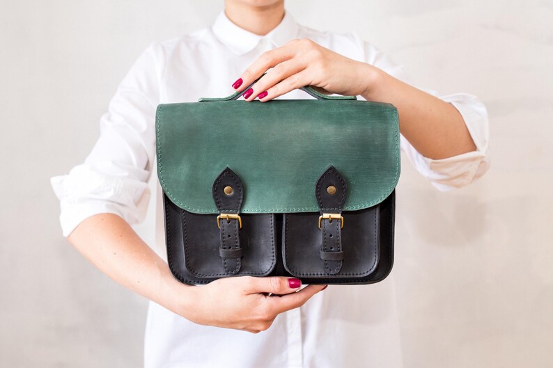 May include: A two-tone leather satchel with a green flap and black body. The bag features two front pockets, gold-coloured buckles, and a top handle. The satchel is held up by a person wearing a white shirt.
