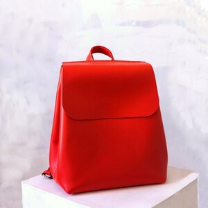 May include: A bright red leather backpack with a flap closure. The backpack has a simple design and is made of high-quality leather.