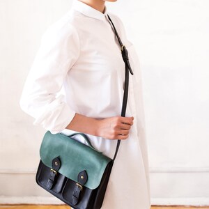 May include: A black and teal leather satchel bag with a flap closure and two front pockets. The bag has a shoulder strap and gold-coloured hardware. The bag is approximately 25 cm wide and 20 cm tall.