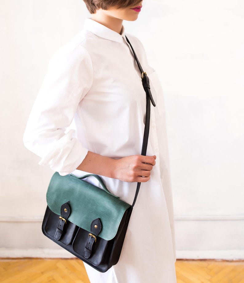 May include: A black and teal leather satchel bag with a flap closure and two front pockets. The bag has a shoulder strap and gold-coloured hardware. The bag is approximately 25 cm wide and 20 cm tall.