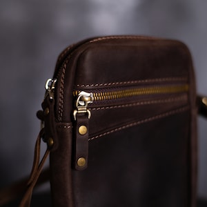 May include: A brown leather crossbody bag with a zipper closure and a gold metal zipper pull. The bag has a strap that can be adjusted to fit different body sizes.