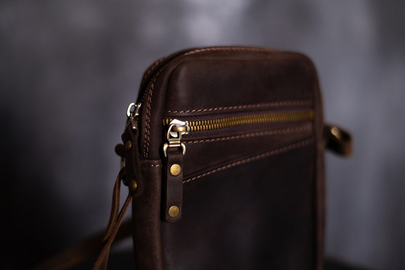 May include: A brown leather crossbody bag with a zipper closure and a gold metal zipper pull. The bag has a strap that can be adjusted to fit different body sizes.