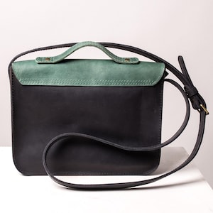 May include: A black leather crossbody bag with a green flap and handle. The bag features a long, adjustable strap and a small handle. The rectangular bag is made of high-quality leather, with a green top section.