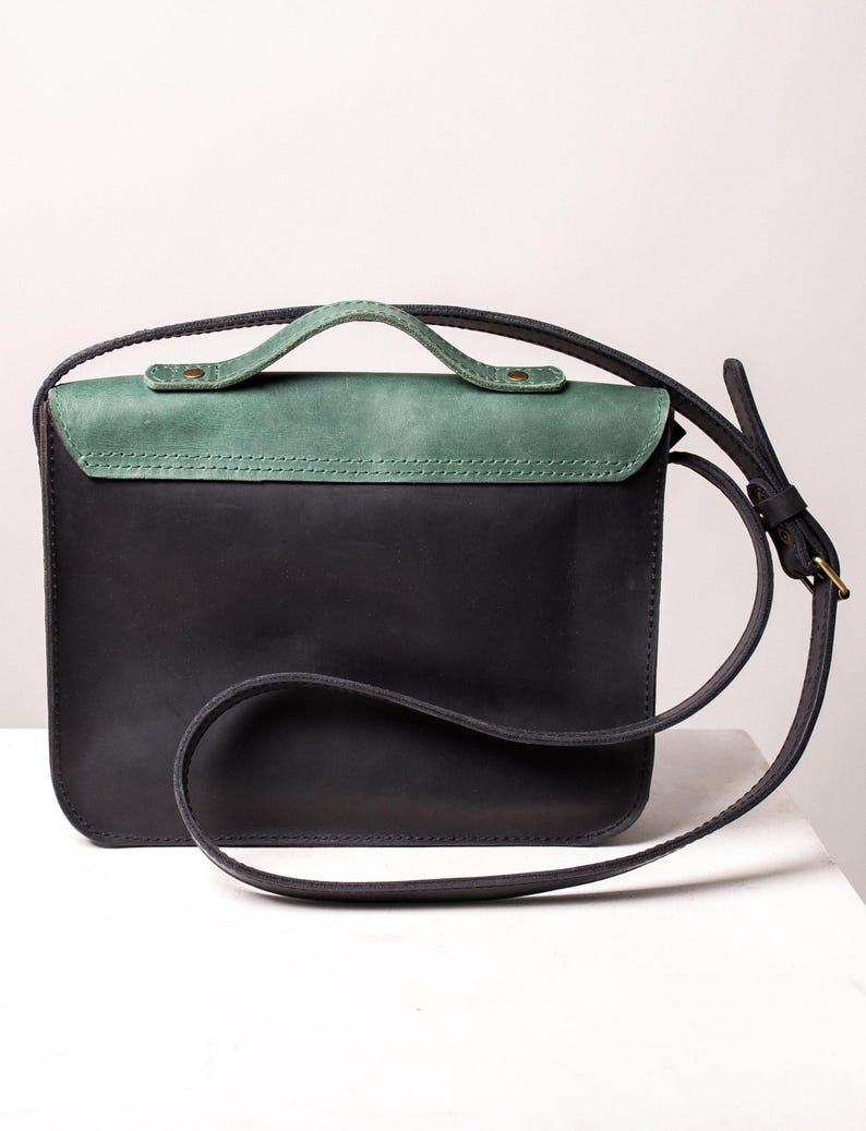May include: A black leather crossbody bag with a green flap and handle. The bag features a long, adjustable strap and a small handle. The rectangular bag is made of high-quality leather, with a green top section.