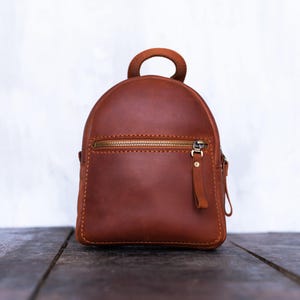 Könnte beinhalten: Ein kleiner, brauner Lederrucksack mit abgerundeter Oberseite und kleinem Griff. Die Tasche hat einen goldfarbenen Reißverschluss und einen passenden Zipper. Der Rucksack steht auf einer dunklen Holzoberfläche.