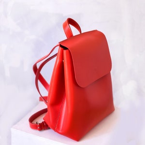 May include: A bright red leather backpack with a flap closure. The backpack has a simple design and is made of high-quality leather.