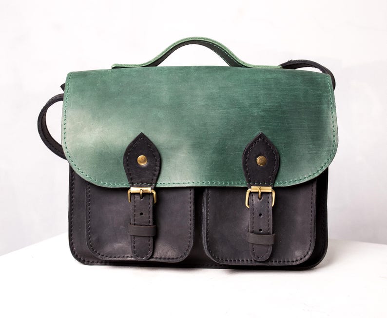 May include: A two-tone leather satchel. The flap is green, the body black. It has two front pockets with brass buckles and a top handle. The shoulder strap is black.