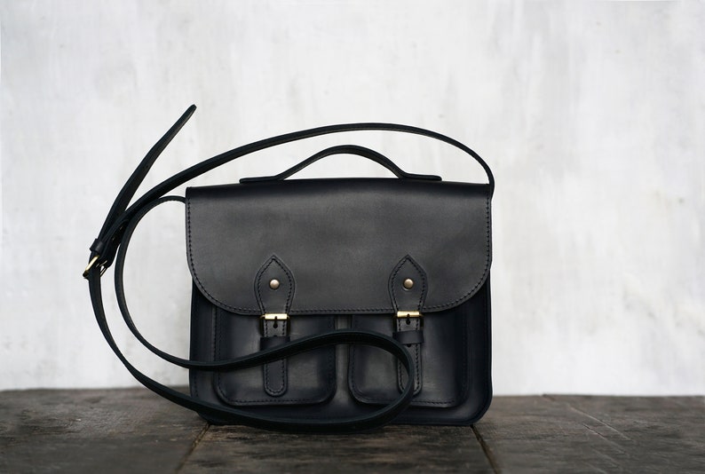 May include: A black leather satchel bag with a flap closure, two front pockets, and a long adjustable shoulder strap. The bag has gold-coloured buckles and hardware. The bag is on a dark wooden surface.