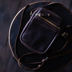 May include: A dark brown leather crossbody bag with a zipper closure and a gold metal zipper pull. The bag has a strap that is adjustable and can be worn over the shoulder or across the body.