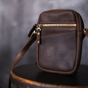 May include: A brown leather crossbody bag with a gold zipper and a flap closure. The bag has a long adjustable strap and a small pocket on the front.