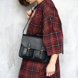 May include: Black leather satchel bag with two front pockets and a flap closure. The bag has gold-coloured buckles and a long shoulder strap. The bag is being held by a person wearing a red and yellow plaid dress.