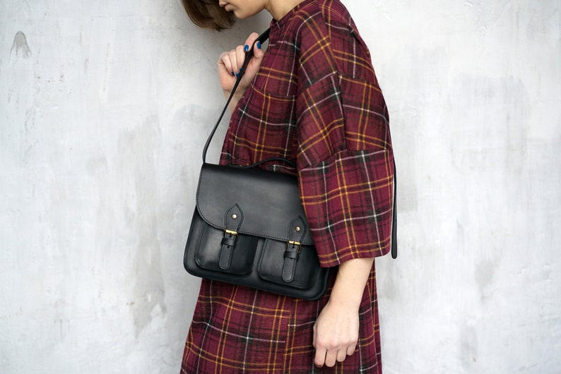 May include: Black leather satchel bag with two front pockets and a flap closure. The bag has gold-coloured buckles and a long shoulder strap. The bag is being held by a person wearing a red and yellow plaid dress.