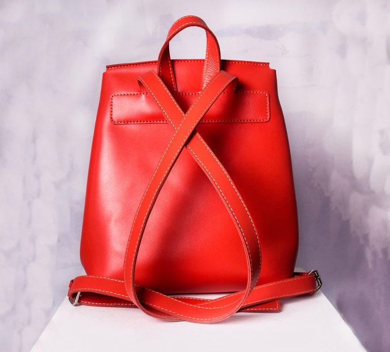 May include: A bright red leather backpack with adjustable straps. The straps are crossed in the back and secured with a buckle.