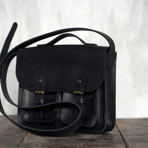 May include: A black leather satchel bag with a flap closure and two front pockets. The bag has a top handle and an adjustable shoulder strap. The hardware is gold-coloured. The bag is on a wooden surface.