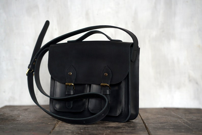 May include: A black leather satchel bag with a flap closure and two front pockets. The bag has a top handle and an adjustable shoulder strap. The hardware is gold-coloured. The bag is on a wooden surface.