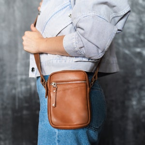 May include: A brown leather crossbody bag with a zipper closure and a silver accent. The bag is worn over the shoulder of a person wearing a light blue denim jacket and jeans.