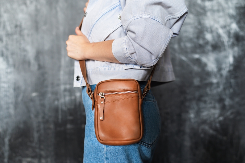 May include: A brown leather crossbody bag with a zipper closure and a silver accent. The bag is worn over the shoulder of a person wearing a light blue denim jacket and jeans.