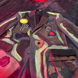 以下が含まれることがあります： 黄色と緑色のペイントアクセントが施されたグレーのブレザー。太陽、文字「P」、ハート、バラのパッチが付いています。ブレザーには迷彩柄のポケットと「BE YOUR BEST」の文字があります。赤とオレンジ色の靴箱が背景にあります。