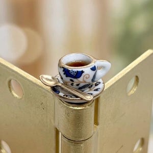 May include: A miniature ceramic teacup and saucer set featuring a white base with blue floral patterns and gold-toned trim. The cup is filled with a dark brown liquid and is accompanied by a small gold-toned metal spoon resting on the saucer.