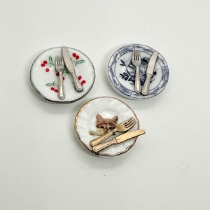 May include: Three miniature plates with silver and gold cutlery. One plate has a red cherry pattern, one has a blue floral pattern, and one has a cat illustration.