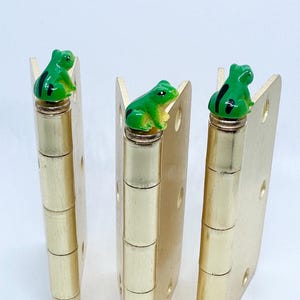 May include: Three gold door hinges with green frog figurines on top. The frogs are facing different directions.
