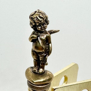 May include: A gold-toned metal bottle stopper with a cherub figurine on top. The cherub is holding a bow and arrow and has wings.