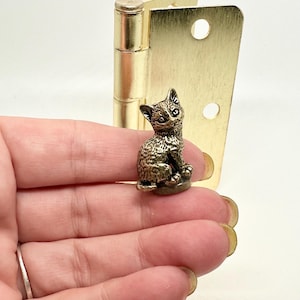 May include: A small, detailed bronze cat figurine sits near a brass hinge. The cat is in a seated position, with intricate detailing on its fur and features. The hinge is a bright gold colour.