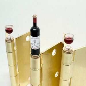 May include: Miniature wine bottle and two wine glasses with red liquid, set on gold-colored hinges. The wine bottle has a label that reads "LOUIS TOKYO" and "Premier Cru". The hinges are set against a white background.