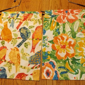 May include: Two decorative kitchen towels. One features colorful birds on branches, the other has a floral design. The towels are white with orange, yellow, blue, and green accents. They are hanging on a black hanger.