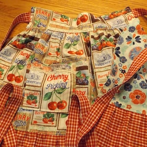 May include: A red and white gingham apron with a patchwork design featuring various berry-themed illustrations. The apron has a large pocket with the text "Cherry Picking" and a ruffled bottom.