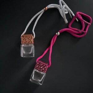 May include: Two glass bottles with decorative metal tops and strings for hanging. The bottle on the left has a white string and the bottle on the right has a pink string.