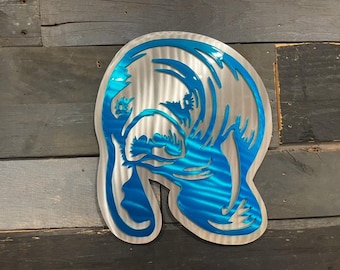 May include: A blue metal wall art sculpture of a manatee. The sculpture is made of brushed metal with a blue finish. The manatee is facing to the right and has a detailed design.