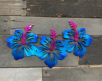 May include: Three metal hibiscus flowers in shades of blue with pink stamens. The flowers are mounted on a weathered wood background, creating a tropical theme. Each flower has detailed petal cutouts.