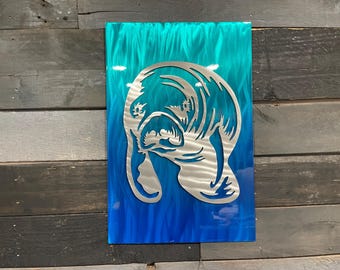 May include: A metal wall art sculpture of a manatee, cut out in a silver color, against a blue and teal gradient background.
