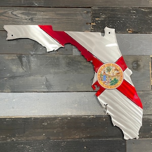 May include: Metal wall art in the shape of the state of Florida with the Florida state flag design. The flag features a red cross on a white background with the state seal in the center. The seal is a circle with a depiction of a woman holding a palm tree and the words "Great Seal of the State of Florida" and "In God We Trust".