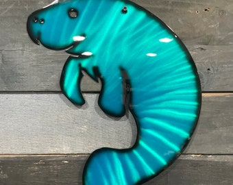 May include: A teal blue metal manatee wall decor with a striped pattern. The manatee is hanging on a wood wall.