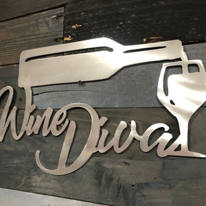 May include: Metal sign with the words "Wine Divas" in a cursive font. A metal silhouette of a wine bottle pouring wine into a glass is above the words.