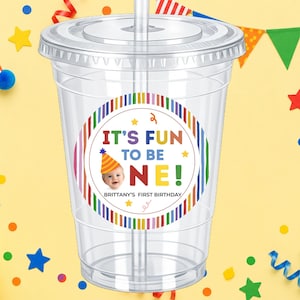 May include: Clear plastic cup with a lid and straw. A colorful sticker on the cup reads "IT'S FUN TO BE ONE!" with a picture of a baby wearing a party hat. The text "BRITTANY'S FIRST BIRTHDAY" is also on the sticker. The background is yellow with party decorations.